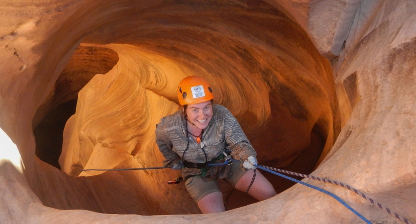 A person wearing safety gear smiles as they rappel into a canyon